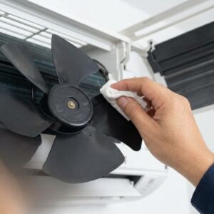 A close-up of an HVAC technician's hand cleaning the fan blade of an air conditioning unit.