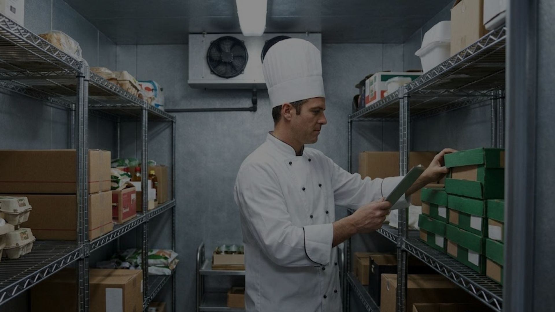 A commercial kitchen chef checks inventory in a well-lit, large walk-in cooler.